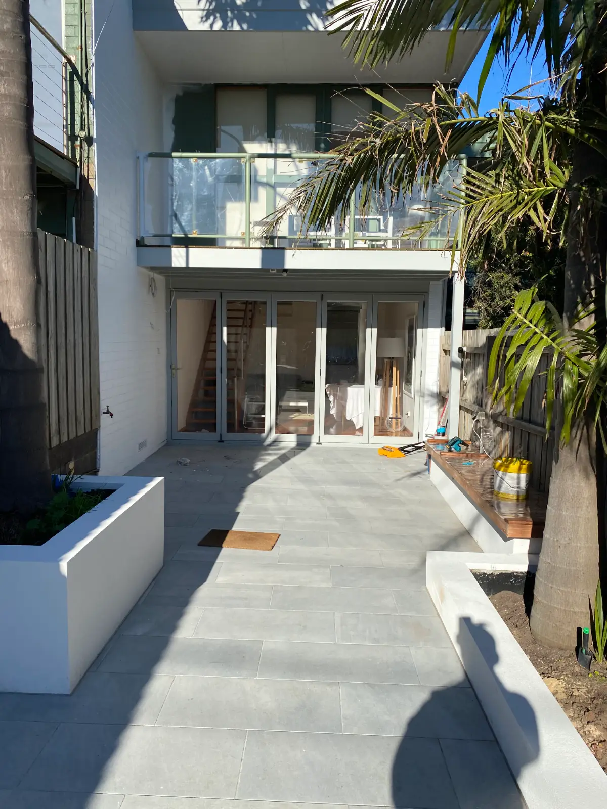 Rear courtyard of a two-storey Mordialloc townhouse during an exterior repaint, white painted brickwork and grey-green joinery below a glass balustrade, with bluestone paving and rendered planters