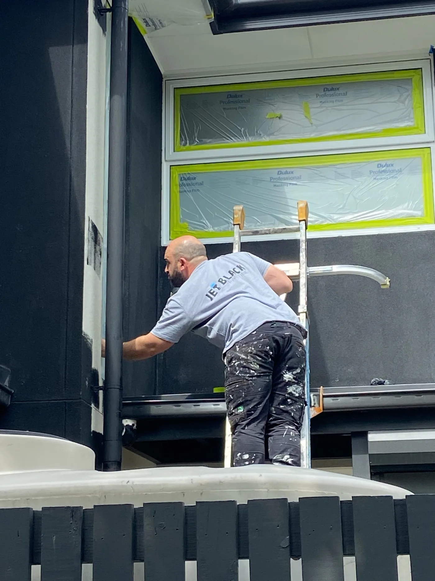 Jimmy Demirci of Jetblack Painting cutting in by hand around a downpipe during an exterior repaint in Mordialloc, with Dulux masking film protecting the windows
