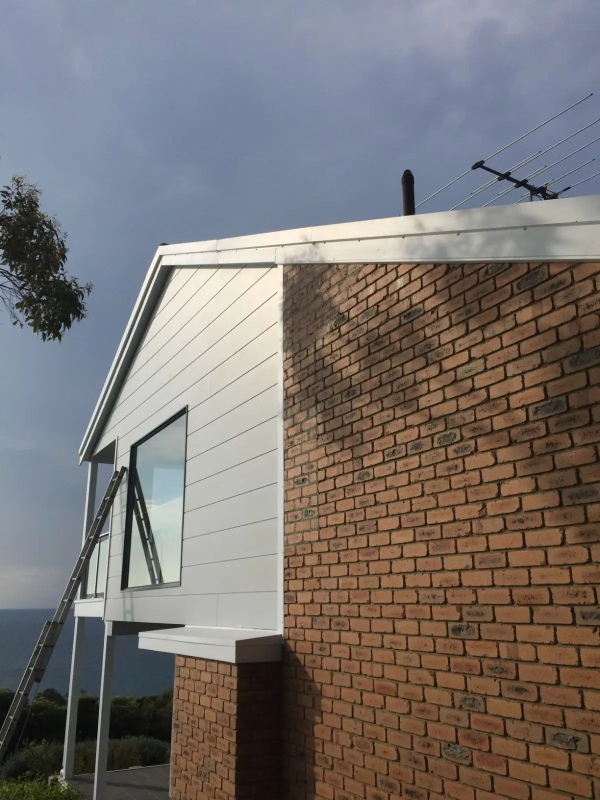 White painted cladding and fascia meeting the original brickwork on the upper gable of a Mount Martha home, with Port Phillip Bay behind