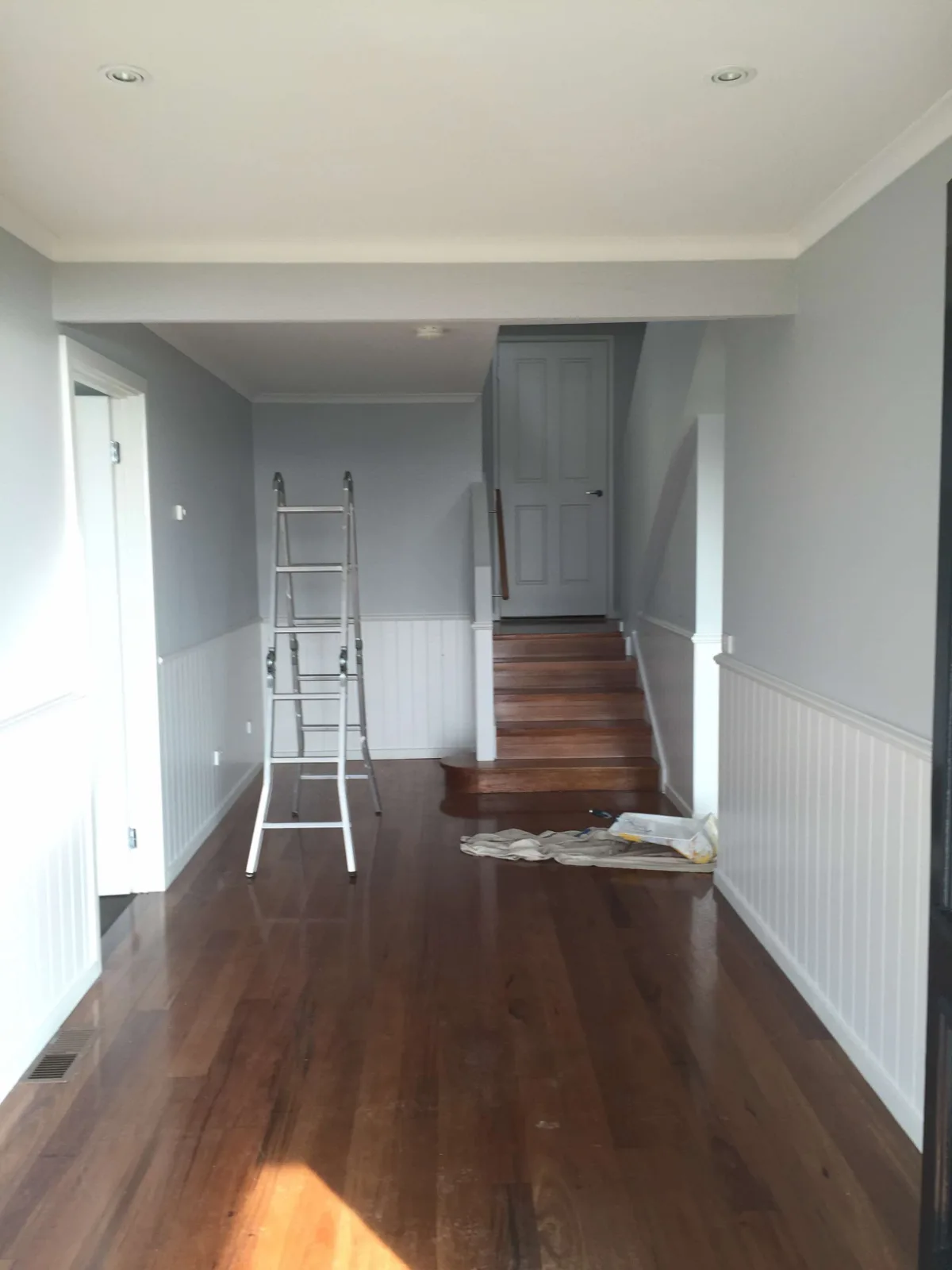 Entry hall of a Mount Martha home mid-repaint, grey walls above white beaded panelling, with timber stairs and floorboards and a ladder and drop sheet in place