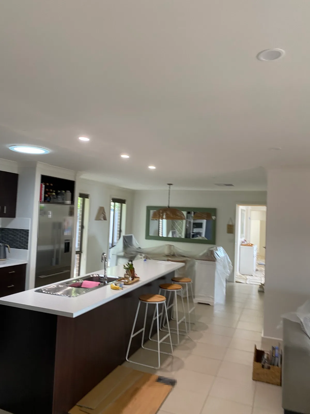 Open-plan kitchen and dining area of a Mount Martha home after an interior repaint, with a freshly painted white ceiling and walls above a dark timber island bench and stools, and furniture covered in plastic