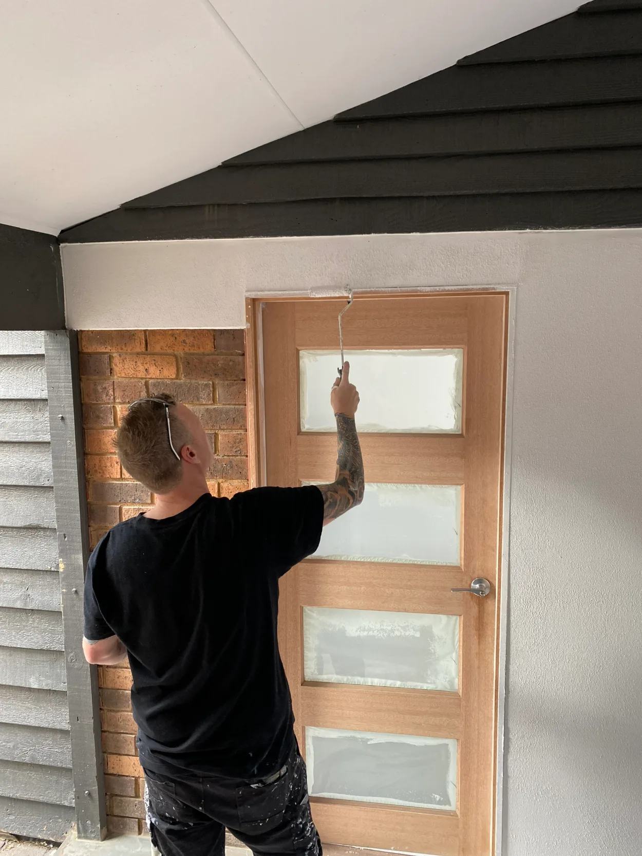 A painter working on the textured wall above a new timber entry door in Narre Warren, the door's glass panels masked, under a gable in black weatherboard