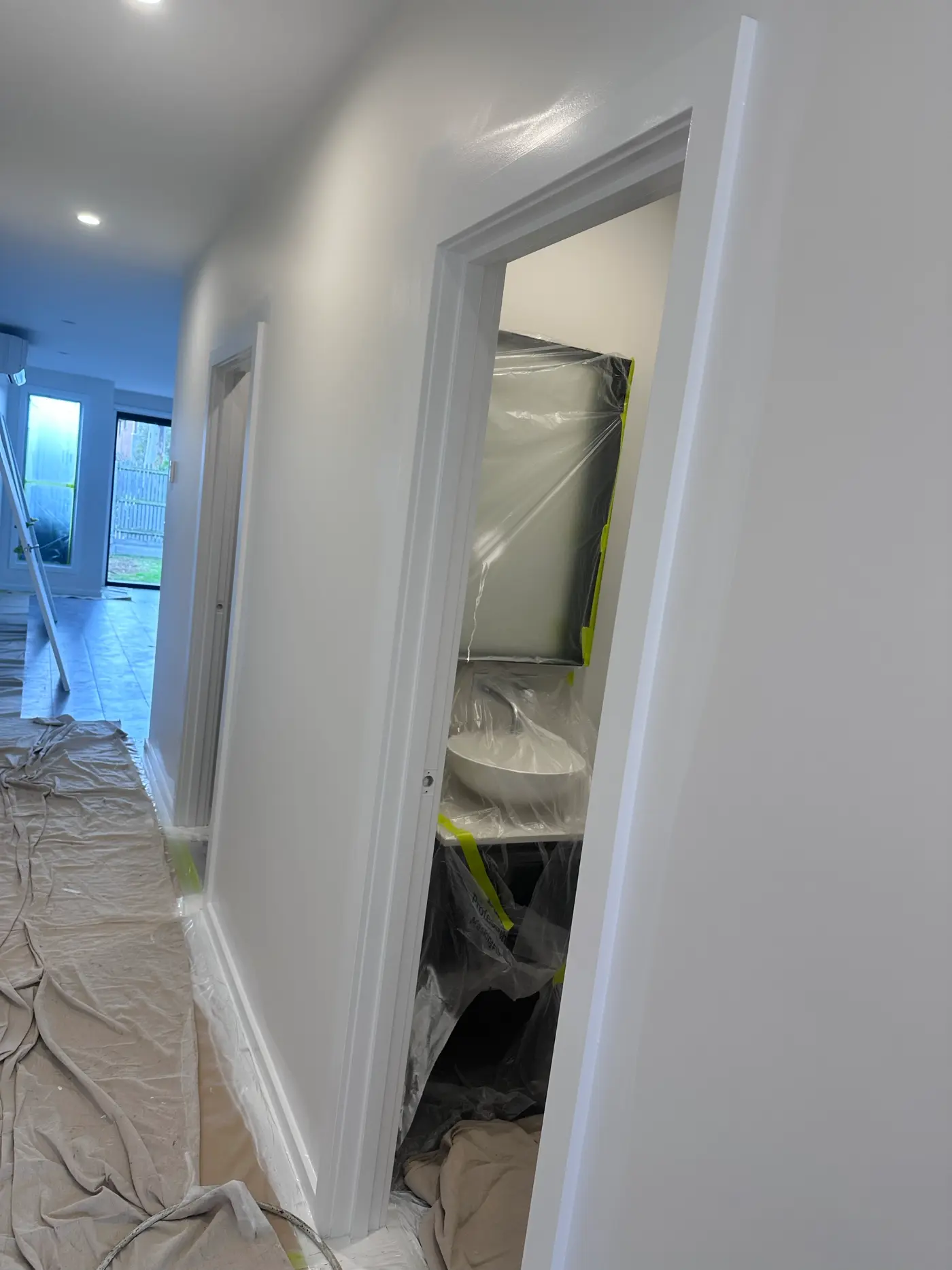 Hallway in a Narre Warren home during repainting, with drop sheets the full length of the floor and the bathroom fixtures wrapped in plastic and masked with tape