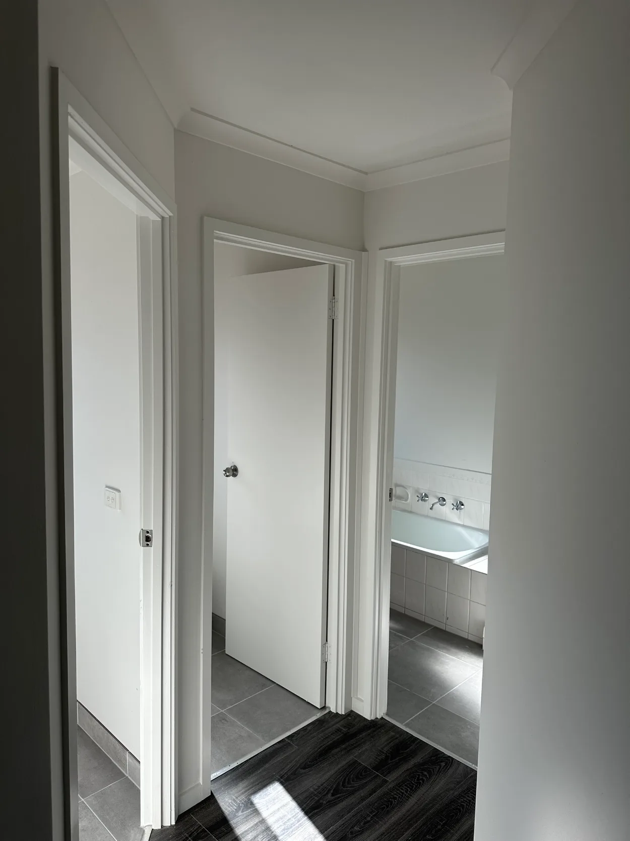 Three doorways off a small landing in a Narre Warren home after repainting, with white walls, ceiling, cornice, architraves and doors above dark timber-look floorboards and grey tile