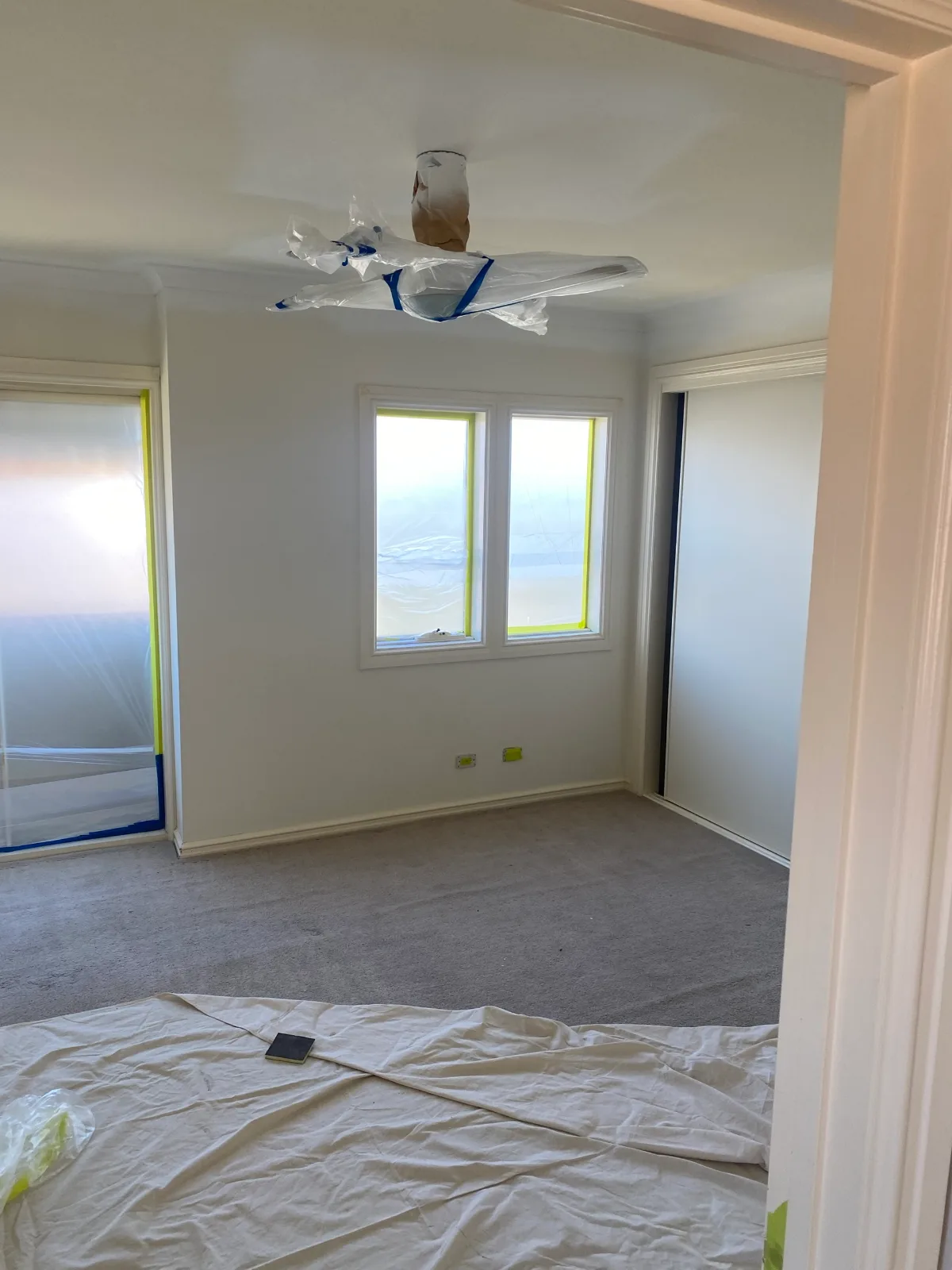 Bedroom in an Oakleigh home during an interior repaint, freshly painted white walls, ceiling and cornice, with the windows and glass door taped and sheeted in plastic, the ceiling light wrapped, and a drop sheet over the carpet