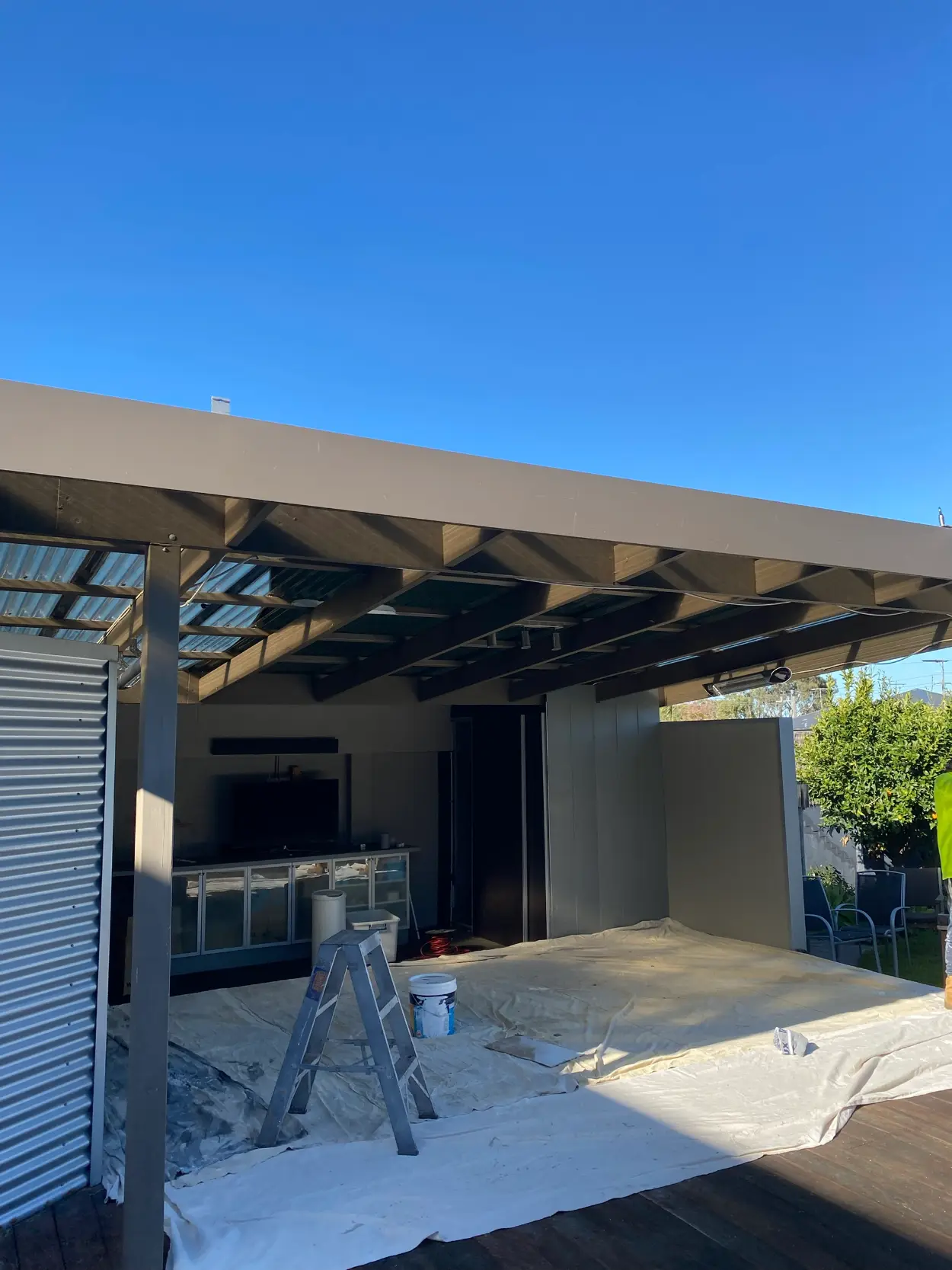 Alfresco area at a Parkdale home during painting, with exposed pergola rafters overhead, charcoal walls, a built-in outdoor kitchen and drop sheets across the deck