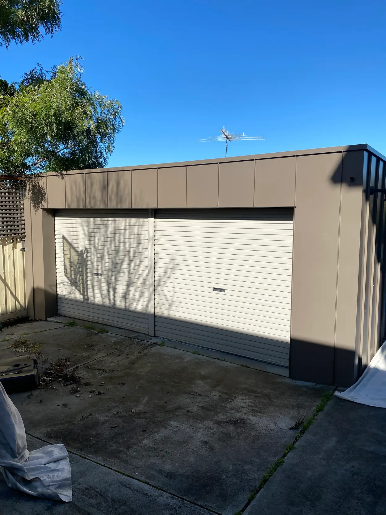 Detached garage at a Parkdale property painted in a warm grey, with a darker charcoal corner trim and two cream roller doors, drop sheets at the edge of the concrete apron