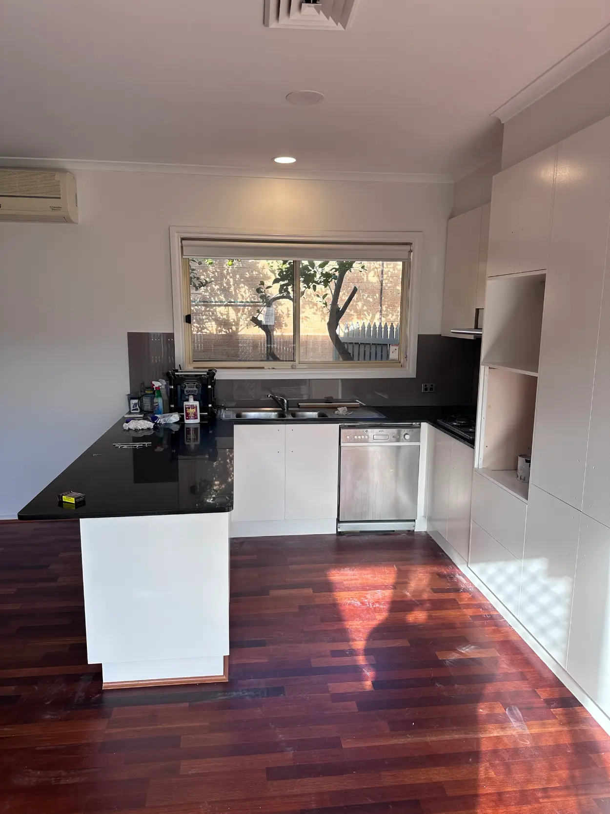 Kitchen in a Parkdale home during work on the joinery, with white cabinetry, a black stone benchtop and grey glass splashback, handles removed and resting on the bench