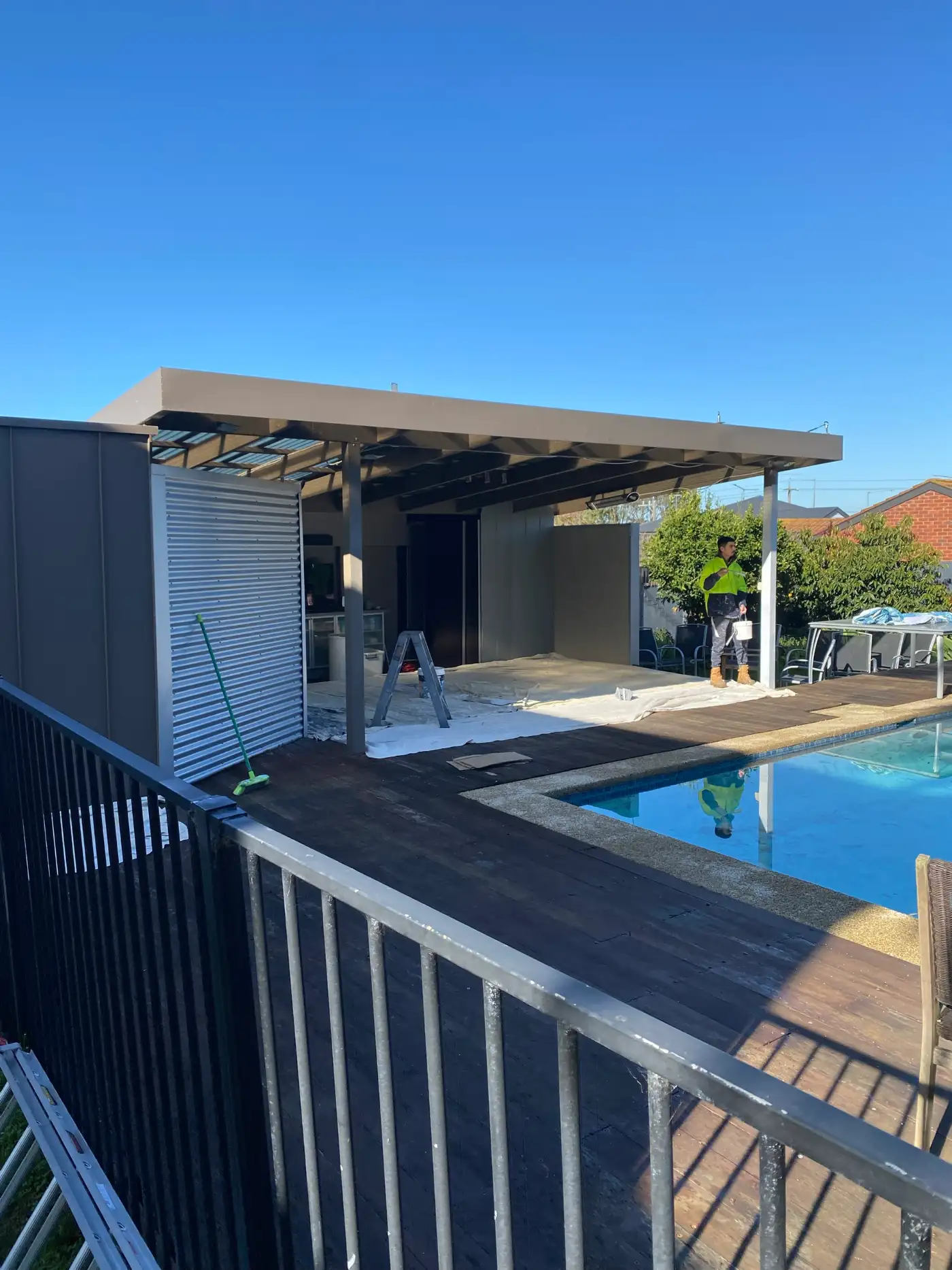 Poolside alfresco at a Parkdale home during painting, with the pergola and rear wall in charcoal, drop sheets across the paved area beneath it, and a painter on the deck beside the pool