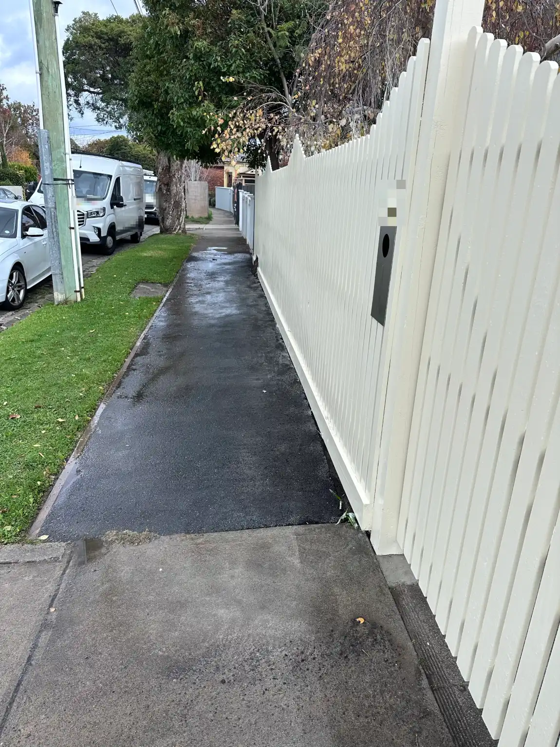 The same white picket fence seen along its full length from the footpath, with a stepped top line and the gate post and letterbox in the foreground