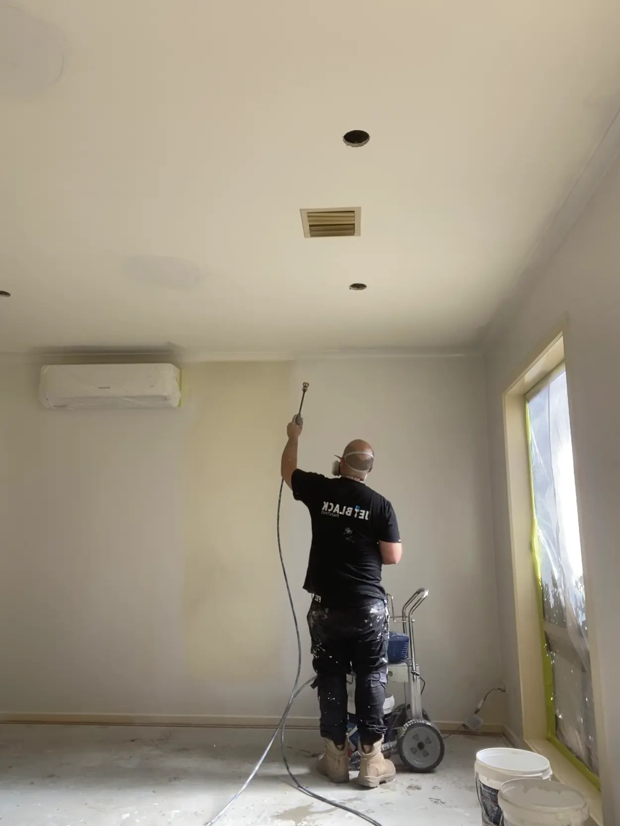 A Jetblack painter in a respirator spraying the walls of a Rowville room with an airless rig, a split-system air conditioner above and the window masked