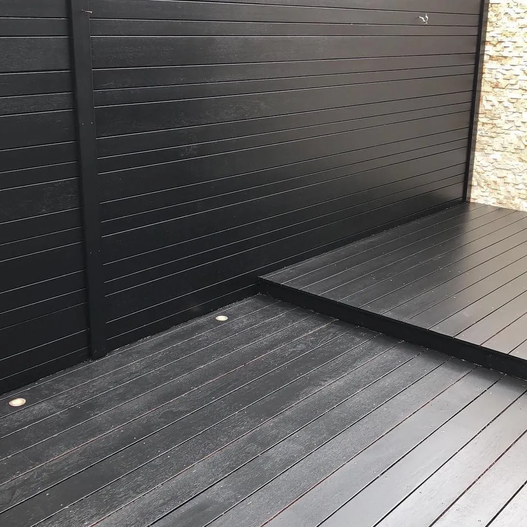 A Sandringham deck stained black, the lower boards and the raised step both, against a black slatted screen and a stacked-stone wall, with small round lights set into the boards