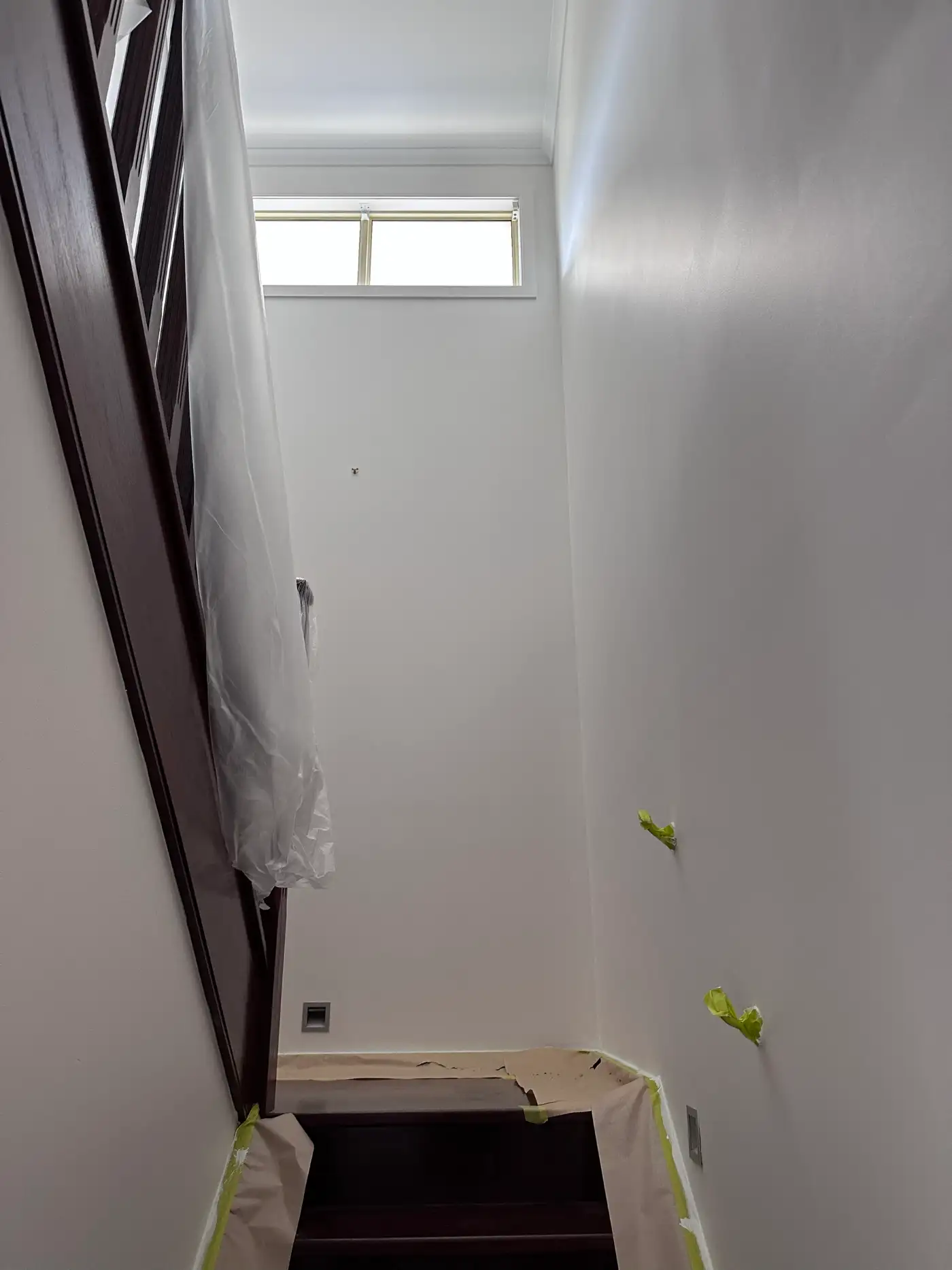 Stairwell in a Sandringham home seen from the lower flight mid-repaint, off-white walls rising to a high window and cornice, with the dark timber handrail wrapped in plastic and the treads covered