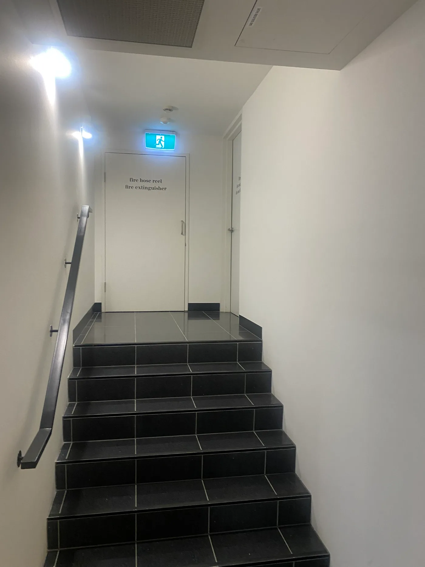 Fire stair in a South Yarra apartment building after repainting, with white walls and ceiling above black tiled treads and a stainless steel handrail