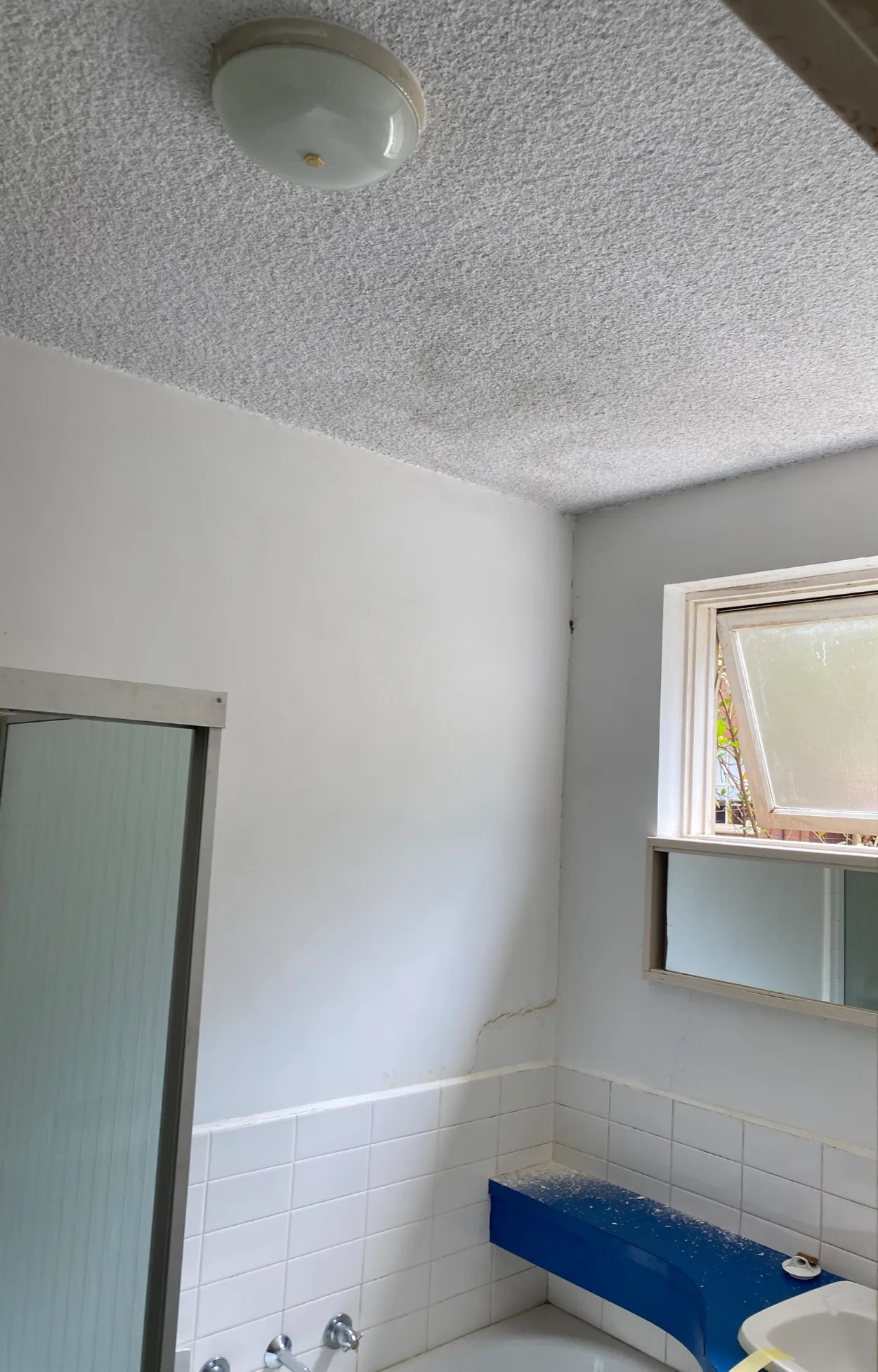 A St Kilda bathroom before painting, with a stippled ceiling showing a dark patch, a crack in the wall above the white tiles, a frosted awning window and a shower screen