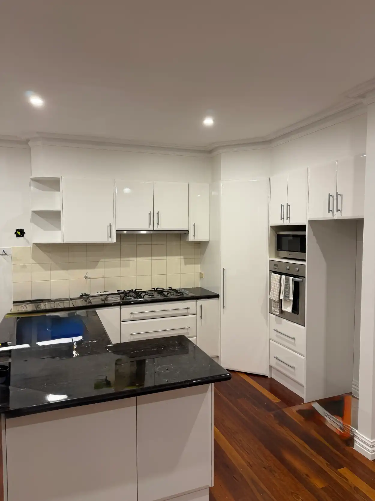 Kitchen in a Stonnington home with its cabinets resurfaced in gloss white, a black granite benchtop still under protective film, a tiled splashback and a tall pantry beside the oven tower