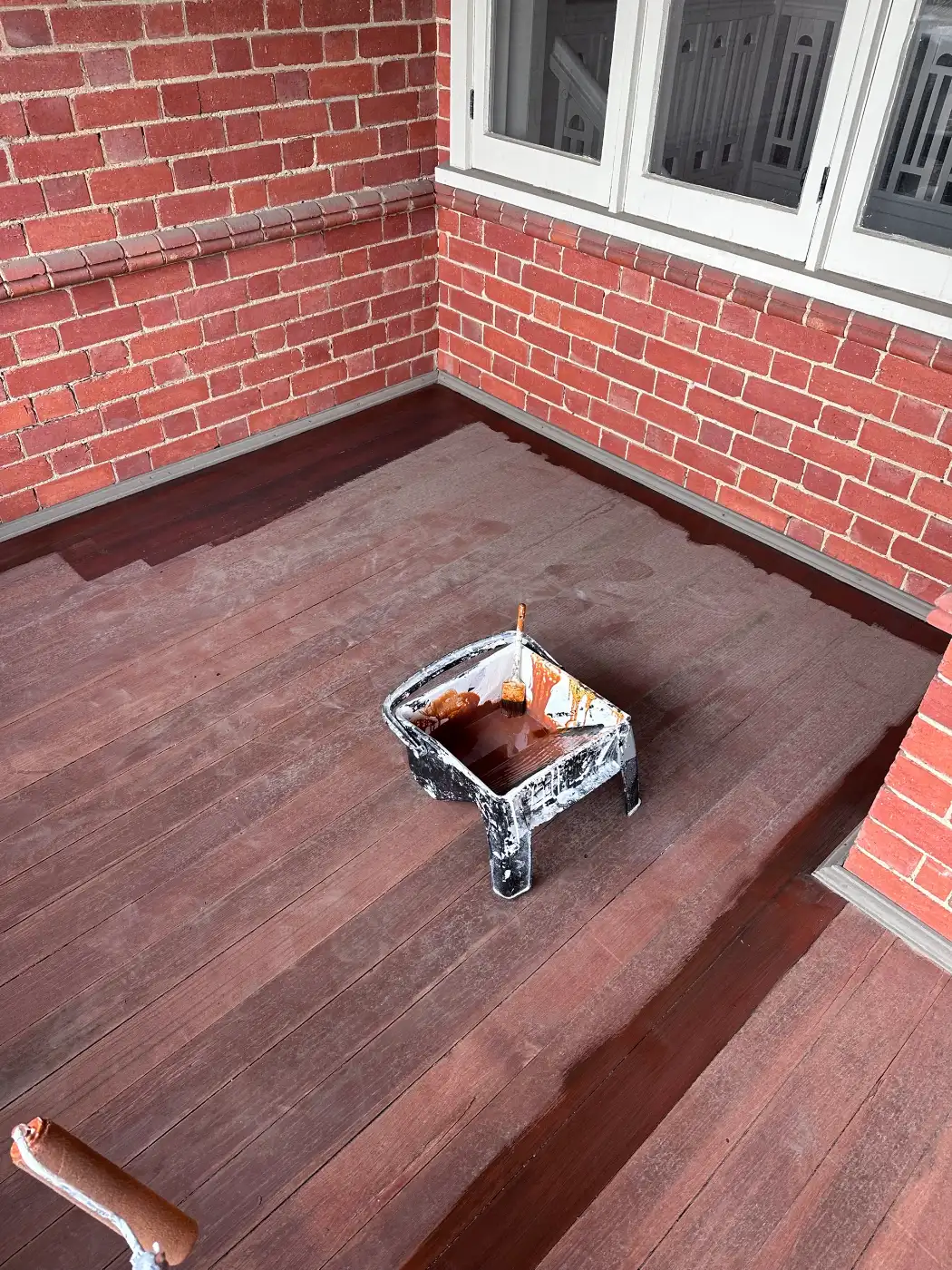 Timber balcony decking on a period Stonnington home part-way through recoating, with the coated boards darker than the bare timber beside them, a tray and brush on the deck