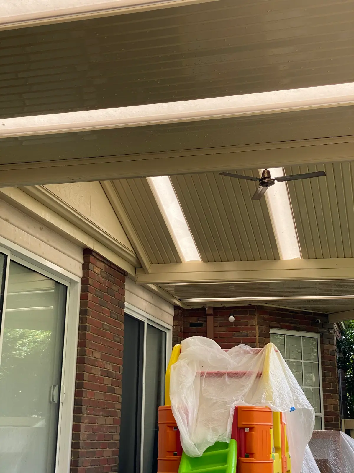 Underside of a metal pergola roof at a Wantirna South brick home before painting, with faded beige sheeting and beams, translucent skylight panels, a ceiling fan, and children's play equipment below wrapped in plastic