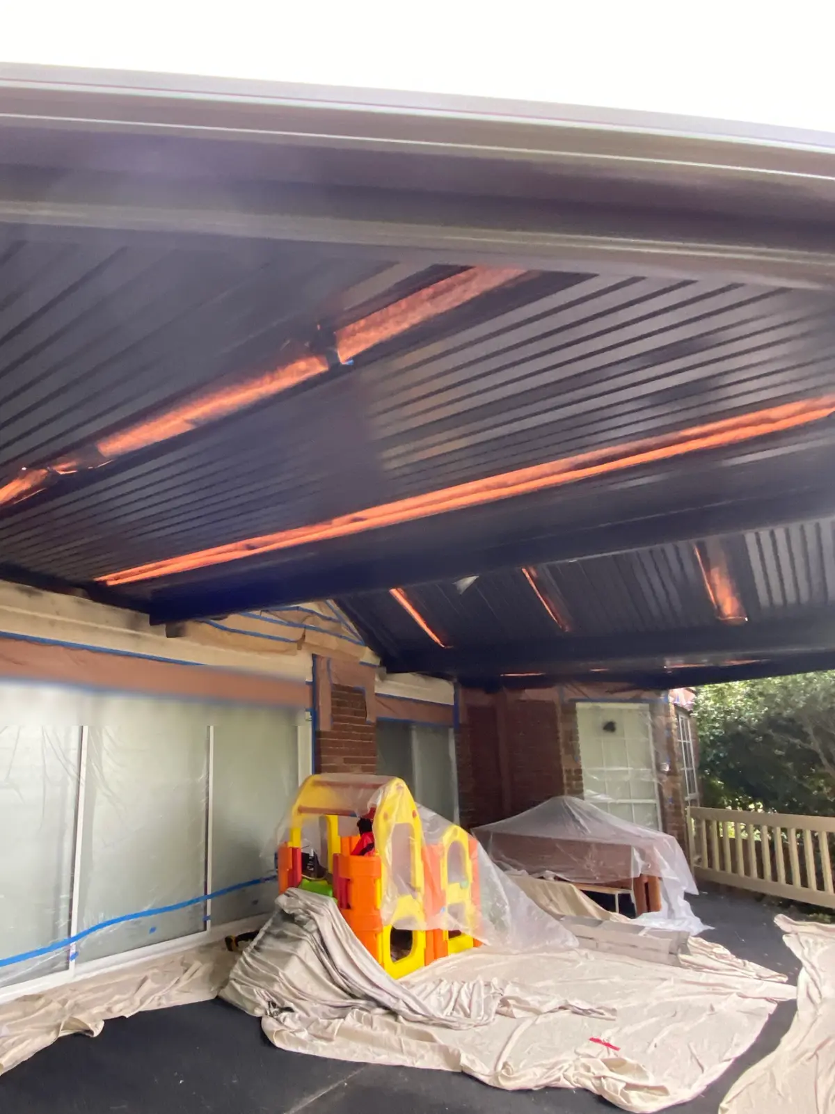 The same Wantirna South pergola mid-job, its roof sheeting and beams now charcoal, with the windows behind masked in plastic film, drop sheets across the paving and the play equipment and outdoor table covered